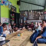 Group of adults seated in a circle on a patterned rug inside a bright green hall for a community workshop or gathering with a projector at the front