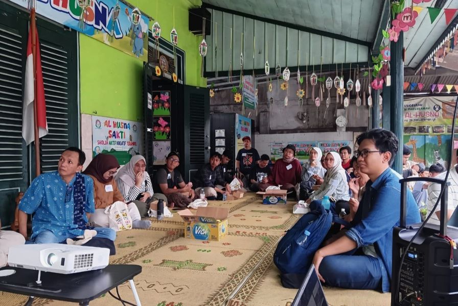 Group of adults seated in a circle on a patterned rug inside a bright green hall for a community workshop or gathering with a projector at the front