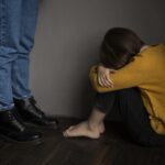 Person in a mustard sweater sits on the floor with head bowed and arms wrapped around their knees beside a standing figure in jeans and black boots