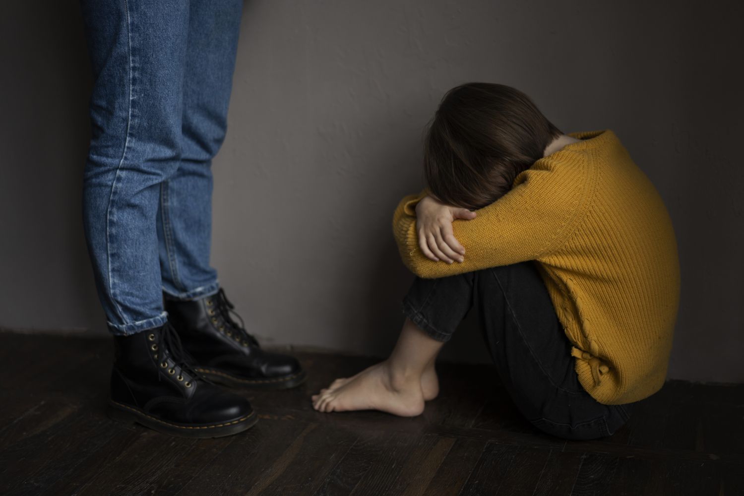Person in a mustard sweater sits on the floor with head bowed and arms wrapped around their knees beside a standing figure in jeans and black boots