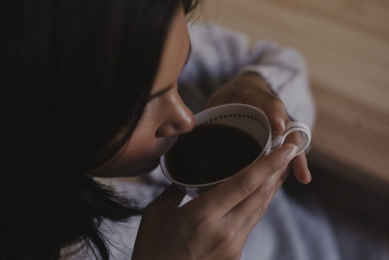 Close-up of a person sipping from a white mug, hands holding the mug near a dark-coated face in a soft, warm setting
