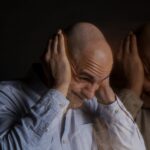 Man with a shaved head in a light blue shirt holds his head in both hands showing stress or frustration with a blurred duplicate beside him