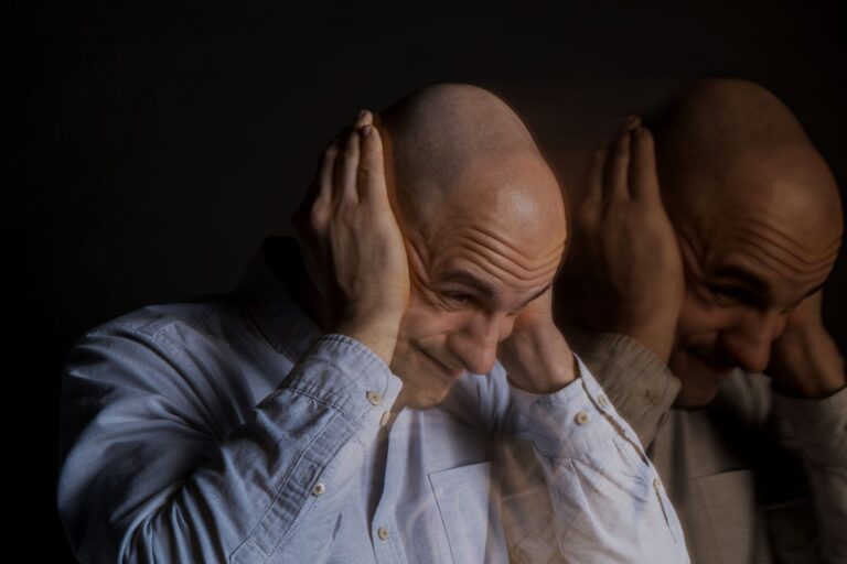 Man with a shaved head in a light blue shirt holds his head in both hands, showing stress or frustration, with a blurred duplicate beside him.