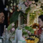 Two older adults in formal attire converse at a table decorated with flowers and fruit with bottles and glasses nearby