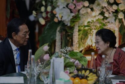 Two older adults in formal attire converse at a table decorated with flowers and fruit, with bottles and glasses nearby.
