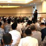 A man in a black robe speaks from a podium on a stage to a large audience seated on the floor in a conference hall many attendees wear patterned batik and head coverings some with masks