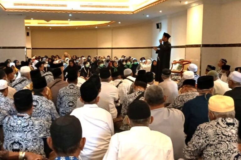 A man in a black robe speaks from a podium on a stage to a large audience seated on the floor in a conference hall; many attendees wear patterned batik and head coverings, some with masks.