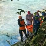 Rescue workers in orange life vests carry an orange tarp along a rocky riverbank beside a blue green river