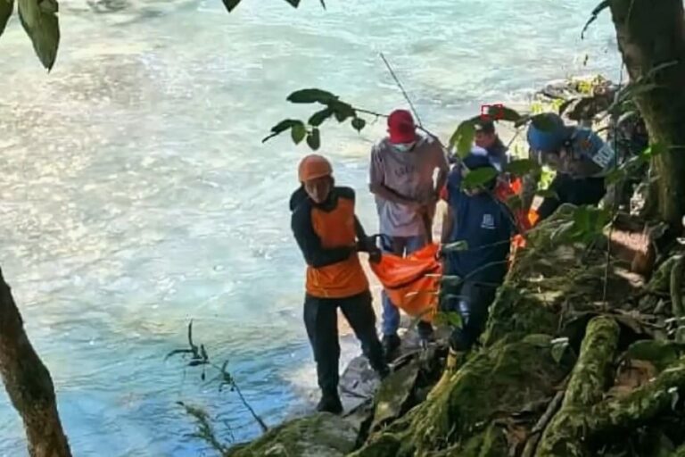 Rescue workers in orange life vests carry an orange tarp along a rocky riverbank beside a blue-green river.