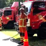 Firefighters in orange turnout gear stand beside red fire trucks with orange cones on a sunny day
