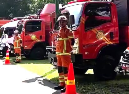 Firefighters in orange turnout gear stand beside red fire trucks with orange cones on a sunny day