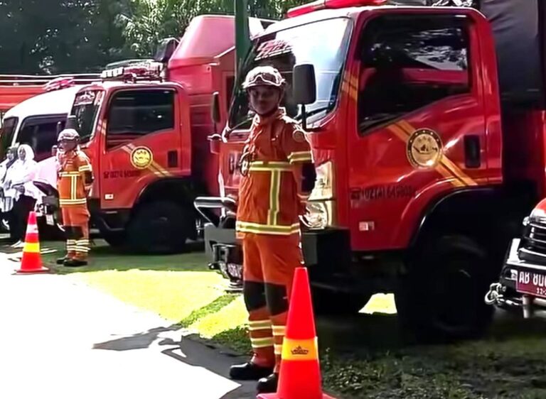 Firefighters in orange turnout gear stand beside red fire trucks with orange cones on a sunny day.