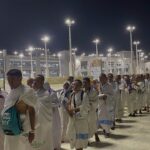 Line of pilgrims wearing white ihram robes standing in a single file outdoors at night under bright floodlights near a large religious complex