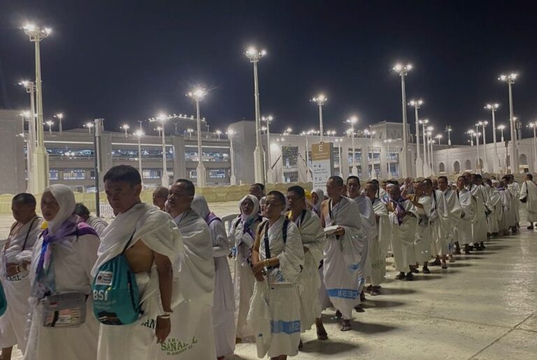 Line of pilgrims wearing white ihram robes standing in a single file outdoors at night, under bright floodlights near a large religious complex.