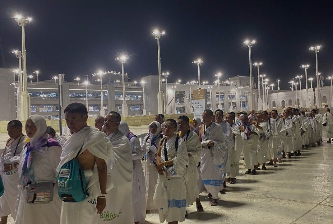 Line of pilgrims wearing white ihram robes standing in a single file outdoors at night under bright floodlights near a large religious complex