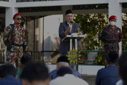 Speaker in suit and traditional cap at podium flanked by two camouflaged soldiers during a formal event audience seated nearby