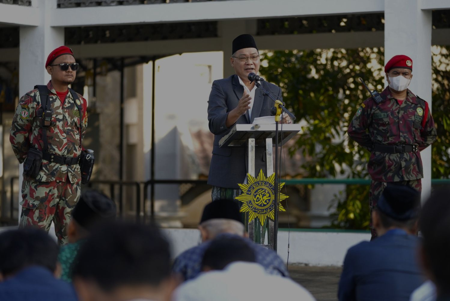 Speaker in suit and traditional cap at podium flanked by two camouflaged soldiers during a formal event audience seated nearby