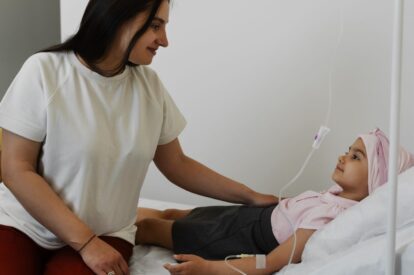 Caregiver sits at the bedside smiling, holding the hand of a young girl in a hospital bed with an IV line