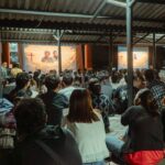 Large crowd seated on a tiled floor under a metal pavilion watching a religious presentation on two screens with crosses and imagery