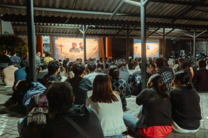 Large crowd seated on a tiled floor under a metal pavilion watching a religious presentation on two screens with crosses and imagery.