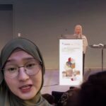 Person wearing a hijab taking a selfie video at a conference with a speaker at a podium in the background and a banner on the lectern