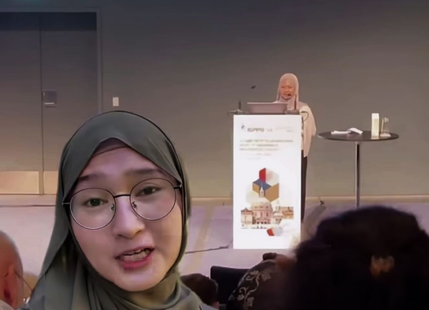 28eriset catrawarta Person wearing a hijab taking a selfie video at a conference with a speaker at a podium in the background and a banner on the lectern