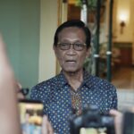 Older man wearing a blue patterned shirt and glasses speaks to a crowd as people take photos with their smartphones nearby