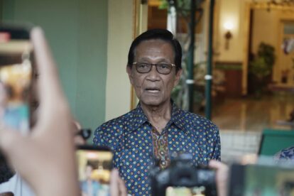 Older man wearing a blue patterned shirt and glasses speaks to a crowd as people take photos with their smartphones nearby.