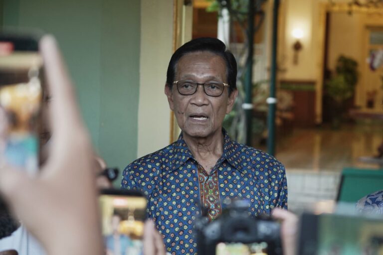 Older man wearing a blue patterned shirt and glasses speaks to a crowd as people take photos with their smartphones nearby.