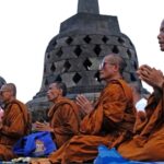 Buddhist monks in orange robes sit in prayer on a platform with large stone stupas in the background