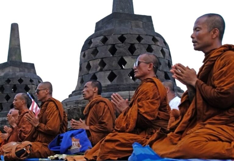 Buddhist monks in orange robes sit in prayer on a platform, with large stone stupas in the background.