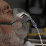 Elderly man wearing a transparent oxygen mask connected to a tube for breathing support in a clinical setting