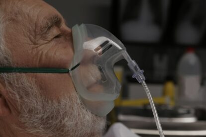 Elderly man wearing a transparent oxygen mask connected to a tube for breathing support in a clinical setting