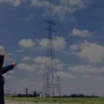 Worker in a white hard hat points at a tall electricity pylon in a grassy field under a blue sky with clouds