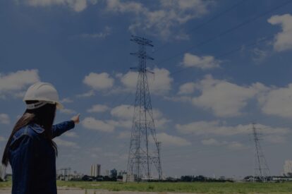 Worker in a white hard hat points at a tall electricity pylon in a grassy field under a blue sky with clouds.