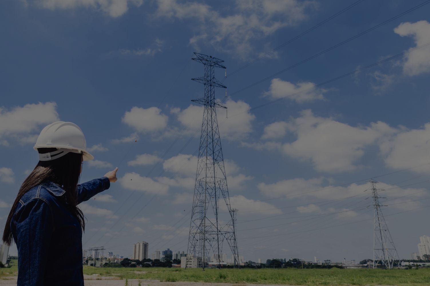 29elistrik catrawarta Worker in a white hard hat points at a tall electricity pylon in a grassy field under a blue sky with clouds