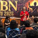 A man in a red shirt speaks into a microphone onstage at an event with a bold banner reading gbn diy 2026 behind him and panelists seated to his right while an audience watches from rows of chairs