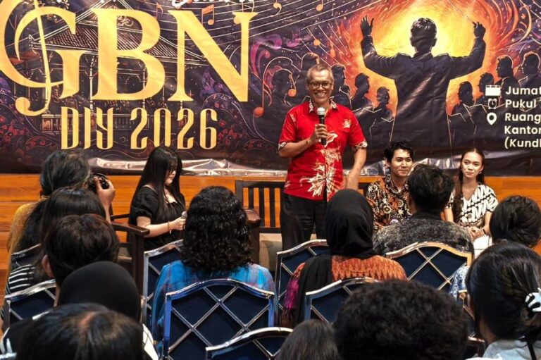 A man in a red shirt speaks into a microphone onstage at an event with a bold banner reading 'GBN DIY 2026' behind him, and panelists seated to his right while an audience watches from rows of chairs.