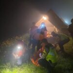 Night time rescue scene with responders in high visibility vests near a lit tent outside flowers and foggy air