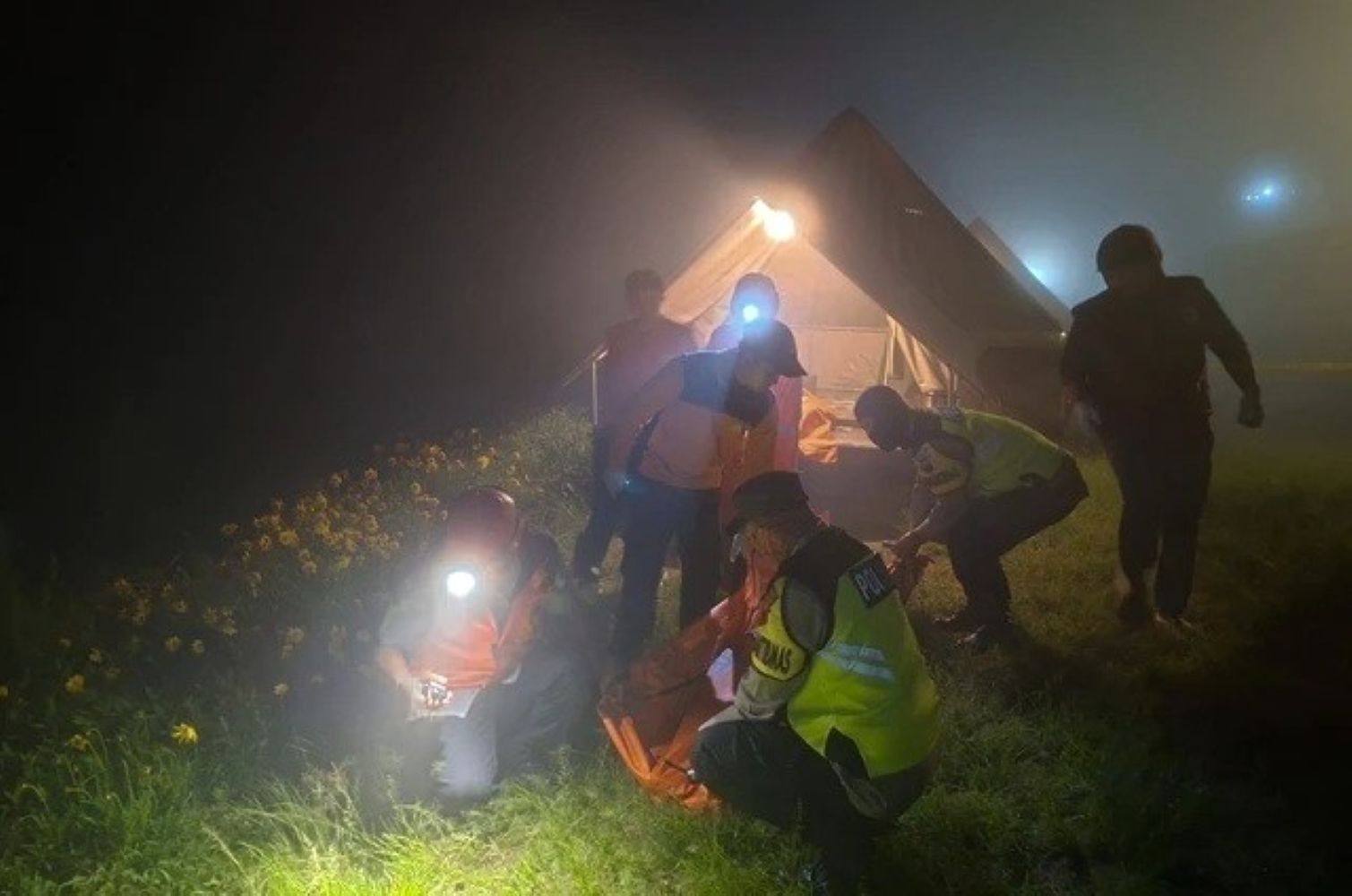 Night time rescue scene with responders in high visibility vests near a lit tent outside flowers and foggy air