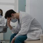 A tired male doctor in a white coat and mask sits on a table head in hand in a clinical room with a coffee cup beside him while a colleague sits in the background wearing a mask