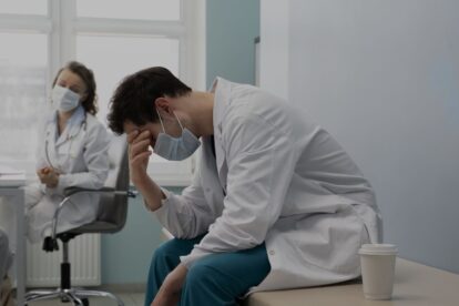 A tired male doctor in a white coat and mask sits on a table, head in hand, in a clinical room with a coffee cup beside him while a colleague sits in the background wearing a mask.