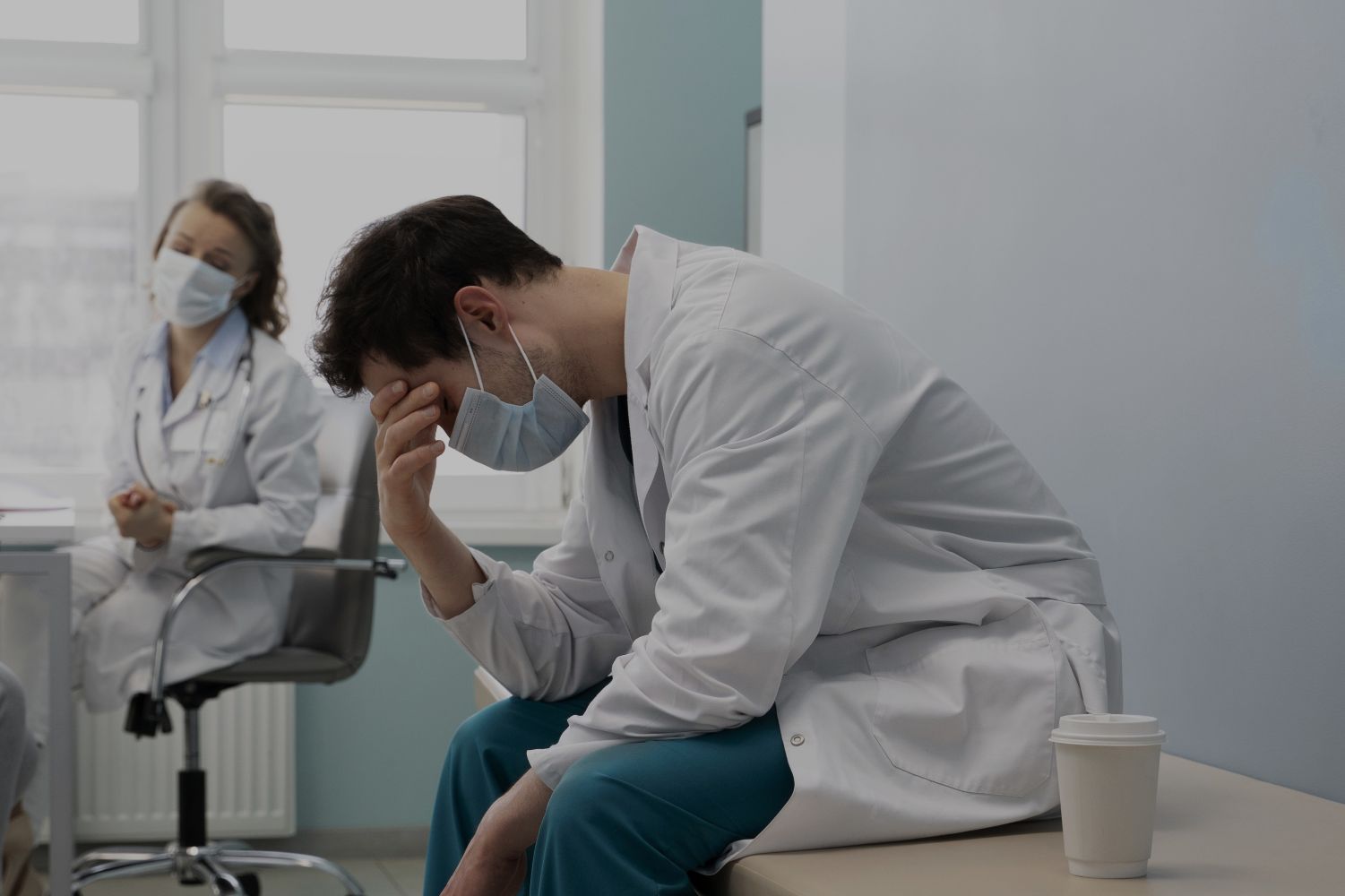 2edokter catrawarta A tired male doctor in a white coat and mask sits on a table head in hand in a clinical room with a coffee cup beside him while a colleague sits in the background wearing a mask