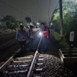 Nighttime rail scene with several people standing on tracks one person in a red safety vest near a signal box