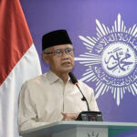Adult man in a beige shirt and black cap delivering a speech at a podium indonesian flag beside him