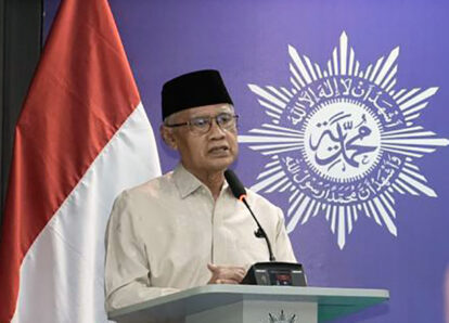 Adult man in a beige shirt and black cap delivering a speech at a podium, Indonesian flag beside him.