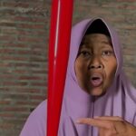 Woman in a lavender hijab speaks to the camera gesturing toward a red vertical pole beside a brick wall background