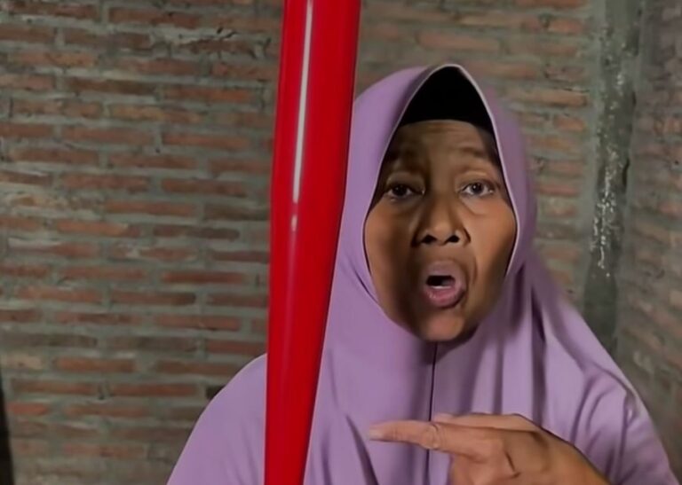 Woman in a lavender hijab speaks to the camera, gesturing toward a red vertical pole beside a brick wall background.