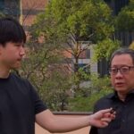 Two men outdoors in a park like area a younger man holds a microphone toward an older man in a black shirt as if conducting an interview