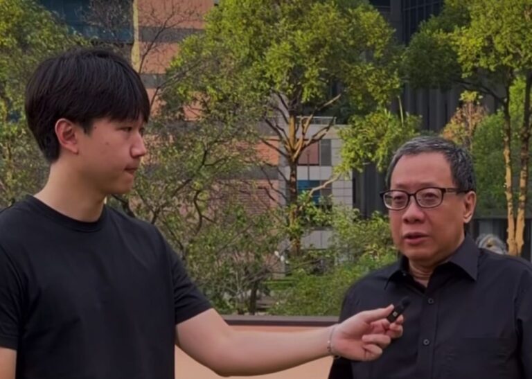 Two men outdoors in a park-like area; a younger man holds a microphone toward an older man in a black shirt, as if conducting an interview.
